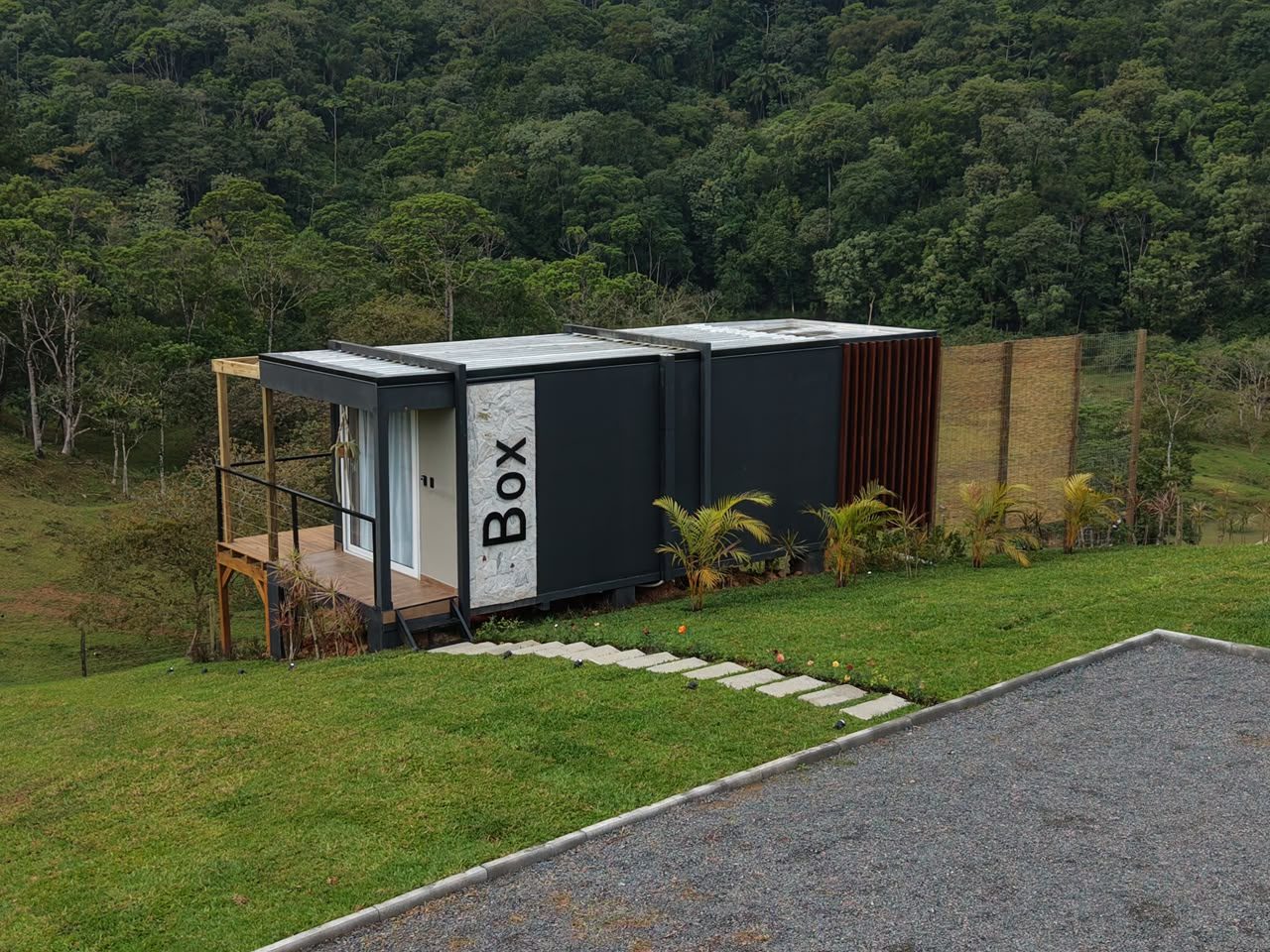 Cabana Box construída pela Construsa em Pomerode, SC: cabana modular preta com painel de pedra e letreiro Box, deck de madeira, pergolado e paisagismo com palmeiras, de frente para a mata