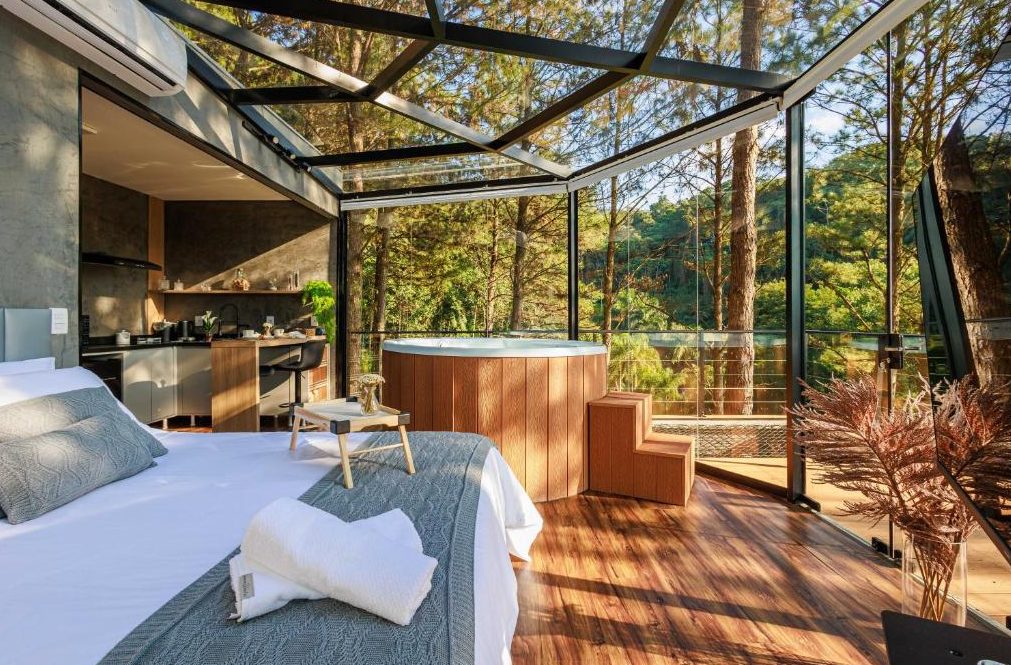 Interior da Cabana Cristallo construída pela Construsa: cabana modular de vidro em meio aos pinheiros, com cama de casal, ofurô de madeira e teto de vidro