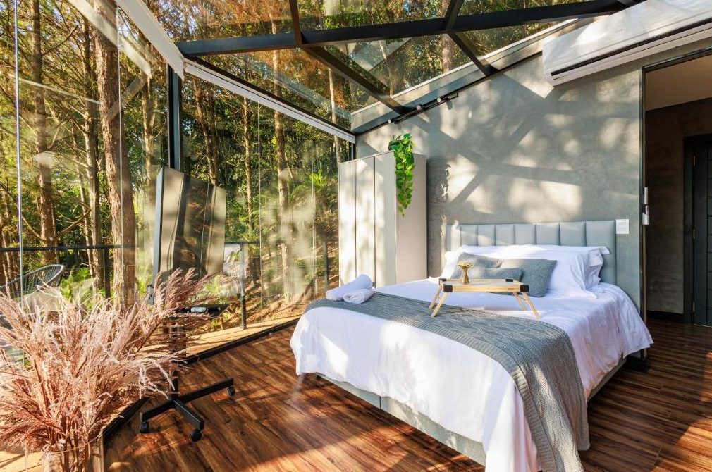 Suíte da Cabana Cristallo com cabeceira estofada, piso de madeira e fechamento em vidro voltado para o pinheiral