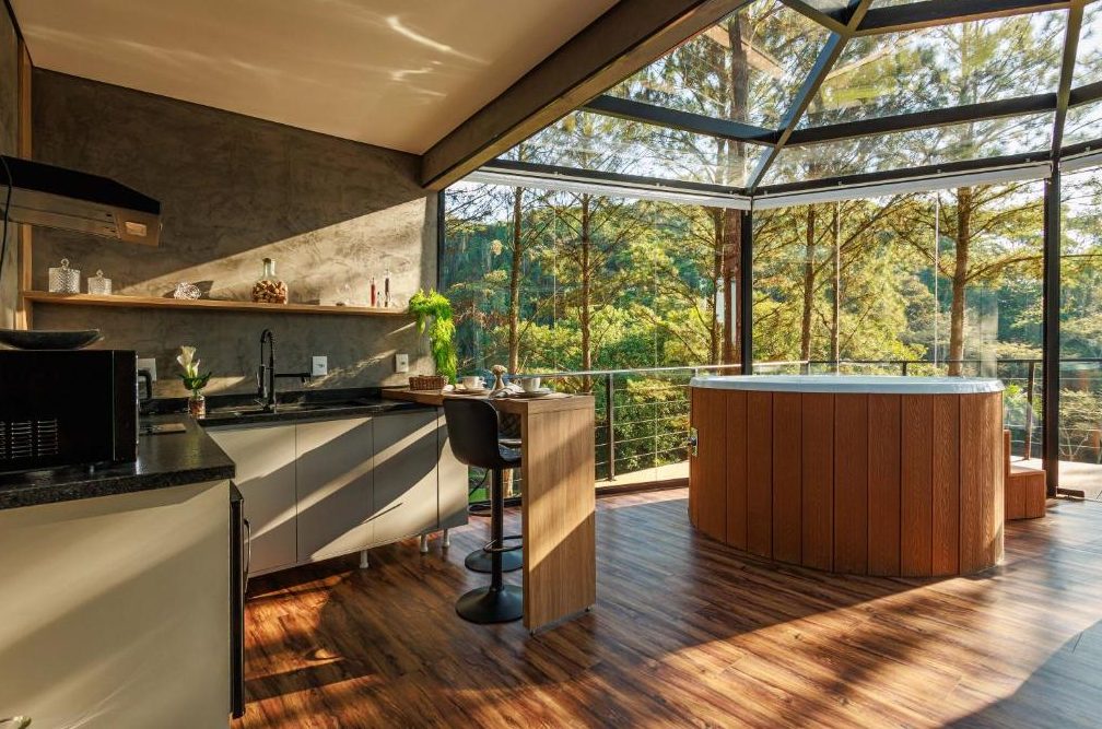 Cozinha da Cabana Cristallo com bancada de granito, mesa alta de madeira e ofurô junto às paredes de vidro