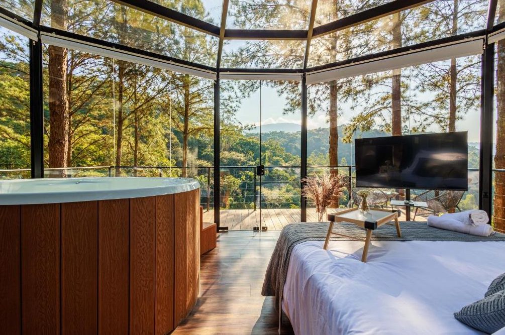 Vista do quarto da Cabana Cristallo para o vale, com ofurô de madeira em primeiro plano e paredes de vidro