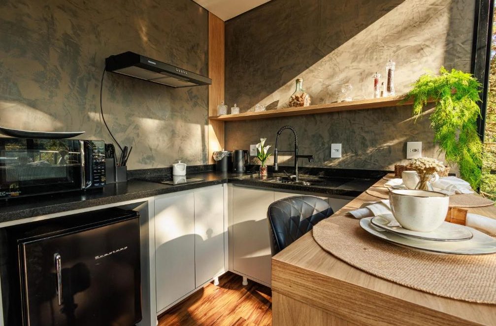 Cozinha compacta da Cabana Cristallo com bancada de granito, parede de cimento queimado e mesa de madeira